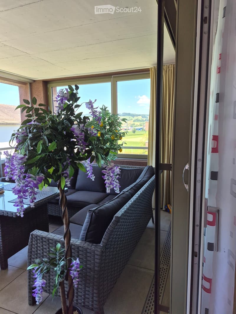 Balcony with a wicker sofa, glass table, and decorative plant with purple flowers, overlooking a scenic countryside view.