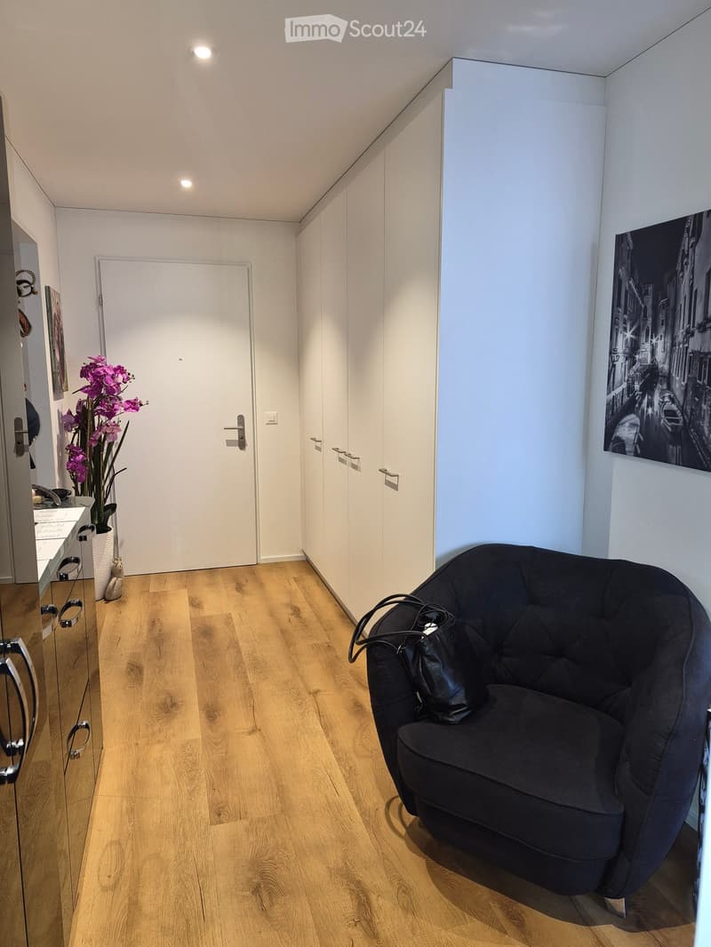 A modern, well-lit hallway with wooden flooring, a black round chair with a handbag, a flower vase with purple flowers, a mirror, and a large picture frame on the wall.