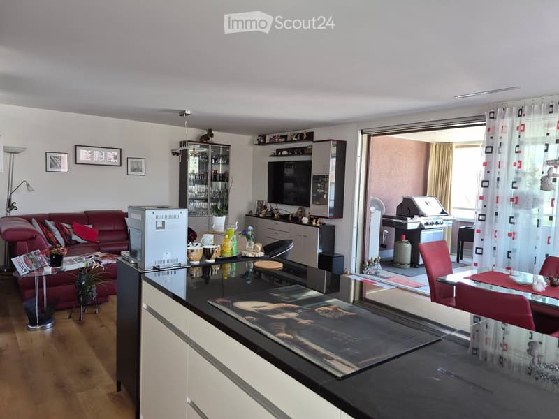 Modern living room with a red sofa, glass coffee table, dining area, TV, and bar area. The room features wooden flooring, large windows with curtains, and a balcony area with a grill and outdoor furniture.