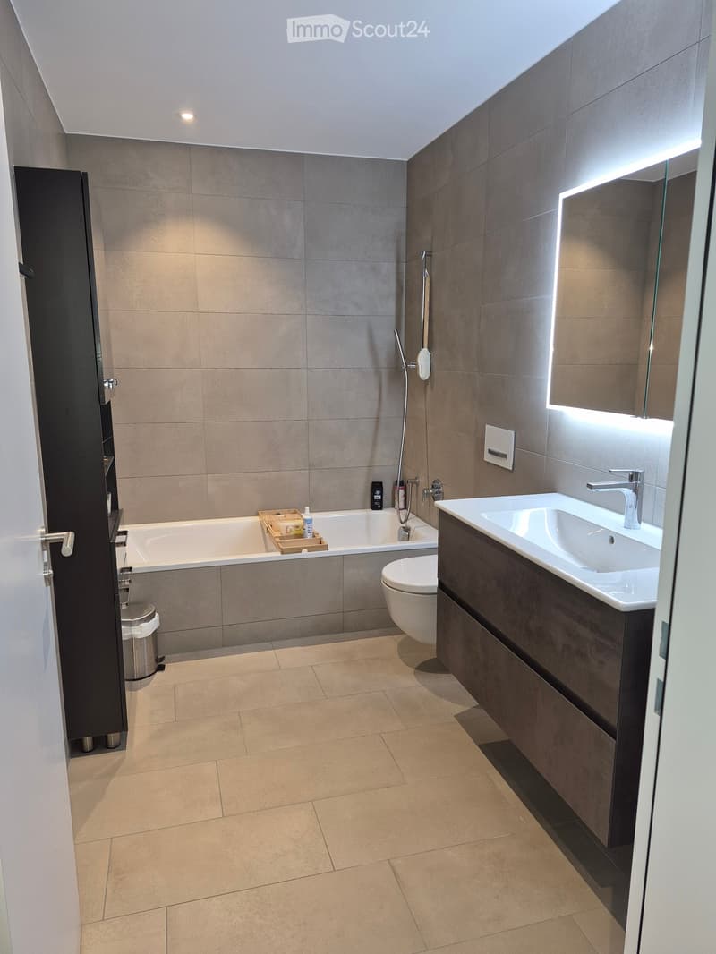 Modern bathroom with a bathtub, toilet, sink, and shower. Features include a large mirror with built-in lighting, a dark wooden vanity, and tiled walls and floor.