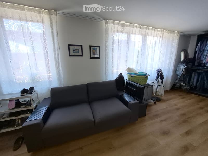 A modern living room with a grey sectional sofa, a small white table with shoes, a television on a stand, and a curtained window. The room features wooden flooring and a small closet with clothes.