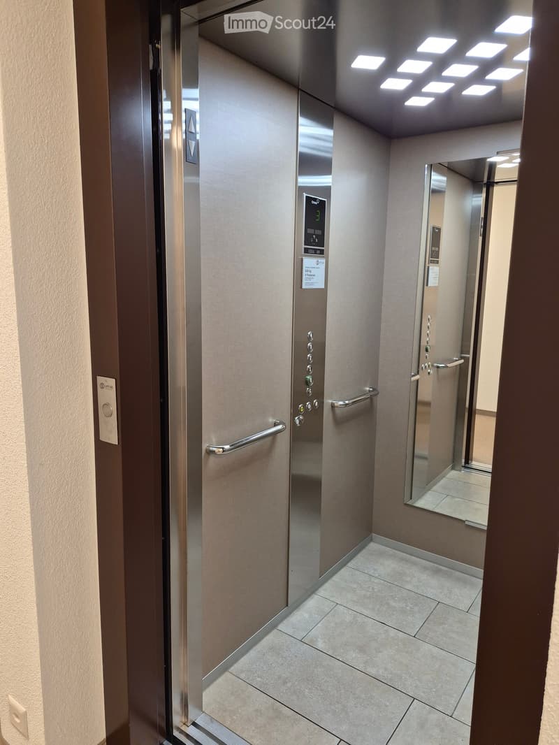 Modern hallway with tiled floor, elevator door, and control panel. The elevator is closed, and the hallway features a clean, contemporary design with neutral tones.