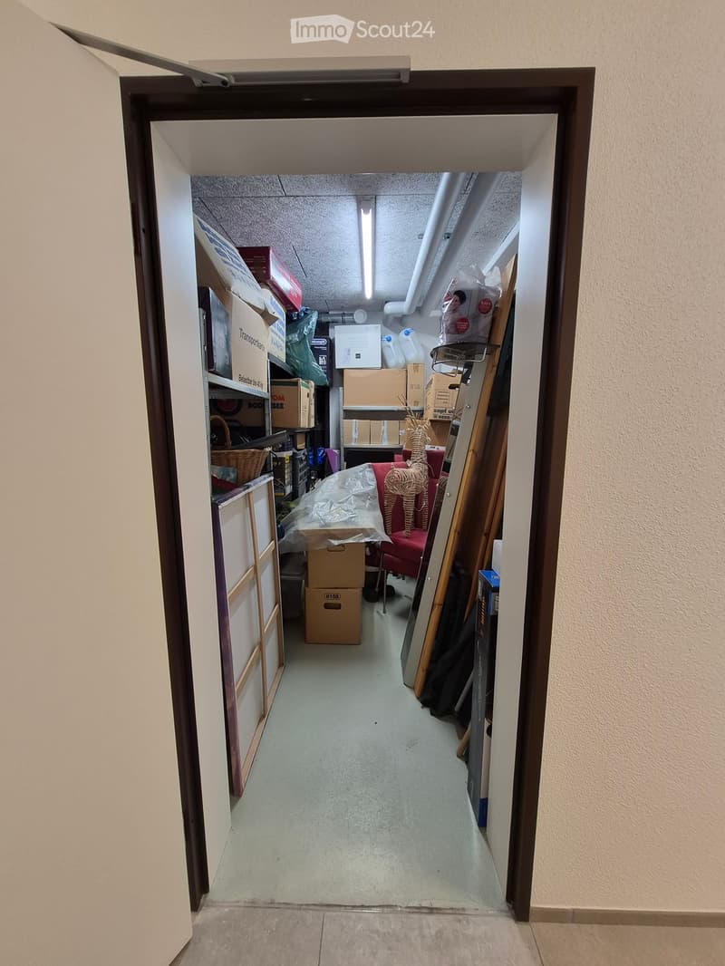A narrow hallway leading into a storage area filled with boxes, furniture, and miscellaneous items. The hallway has beige walls and a concrete floor.