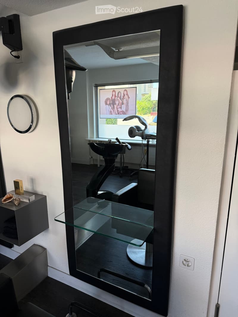 Modern salon with large mirror, glass table, salon chair, and styling station visible in reflection.