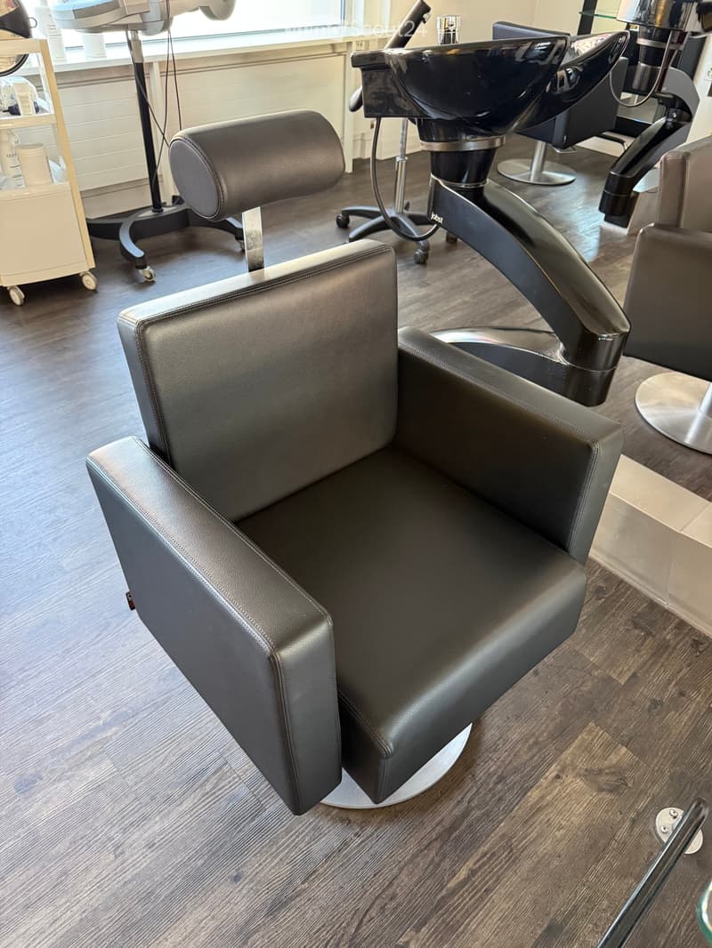 Modern salon with black leather chairs, a sink, and a stool, set on a wooden floor.