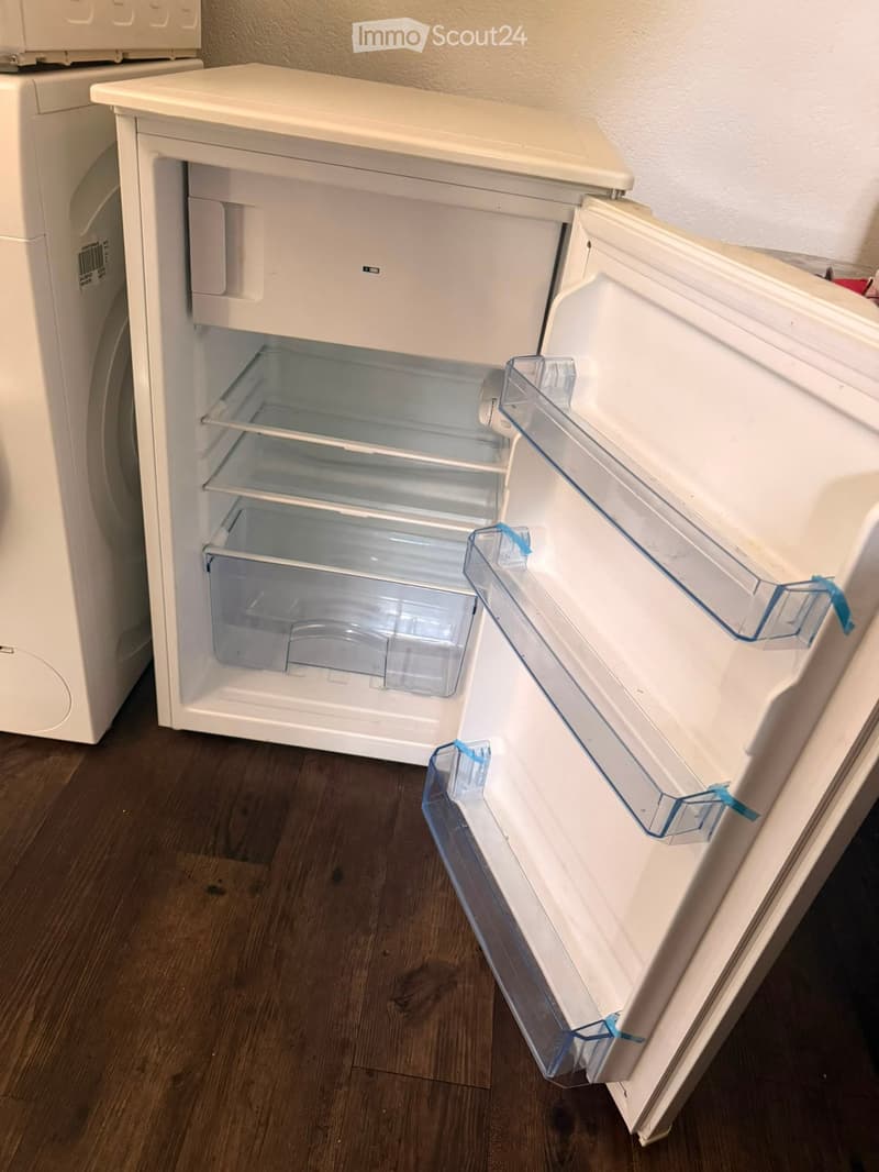 Open refrigerator with empty shelves and drawers, placed on a wooden floor next to a washing machine.