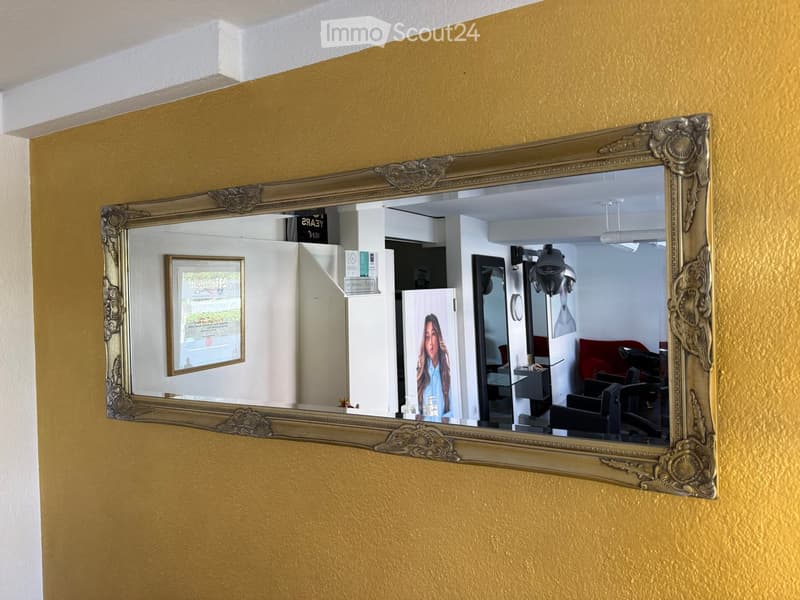 A large ornate mirror reflects a salon or beauty room with chairs, a lamp, a poster, and a cabinet.