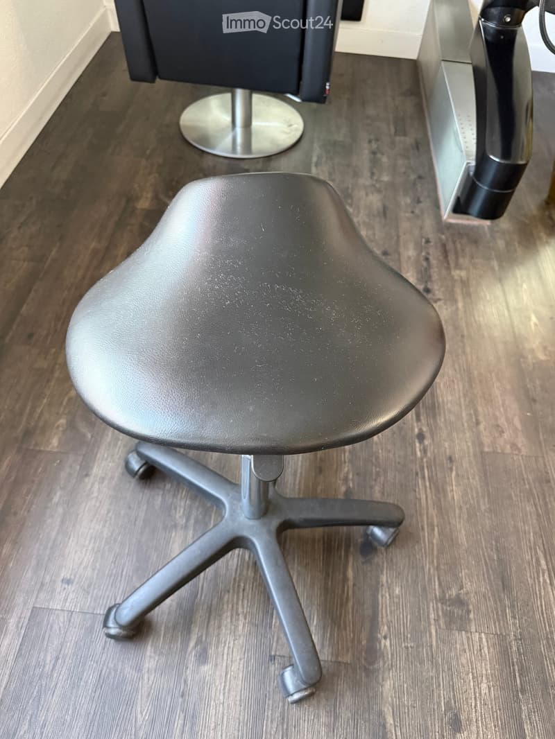 Office chair with a black seat and a grey base, placed on a wooden floor next to a black stool with a silver base.