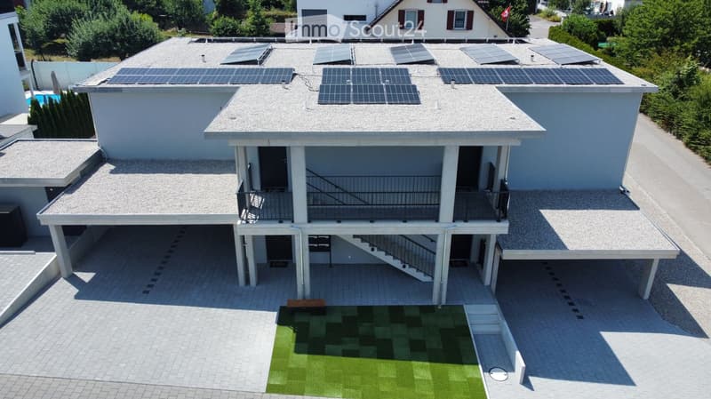 A modern two-story house with a flat roof covered in solar panels, a spacious balcony, and a small artificial grass area on the patio.