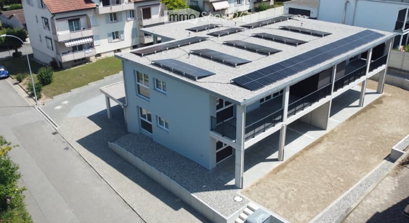 Modern two-story building with solar panels on the roof, balconies, and surrounding greenery.