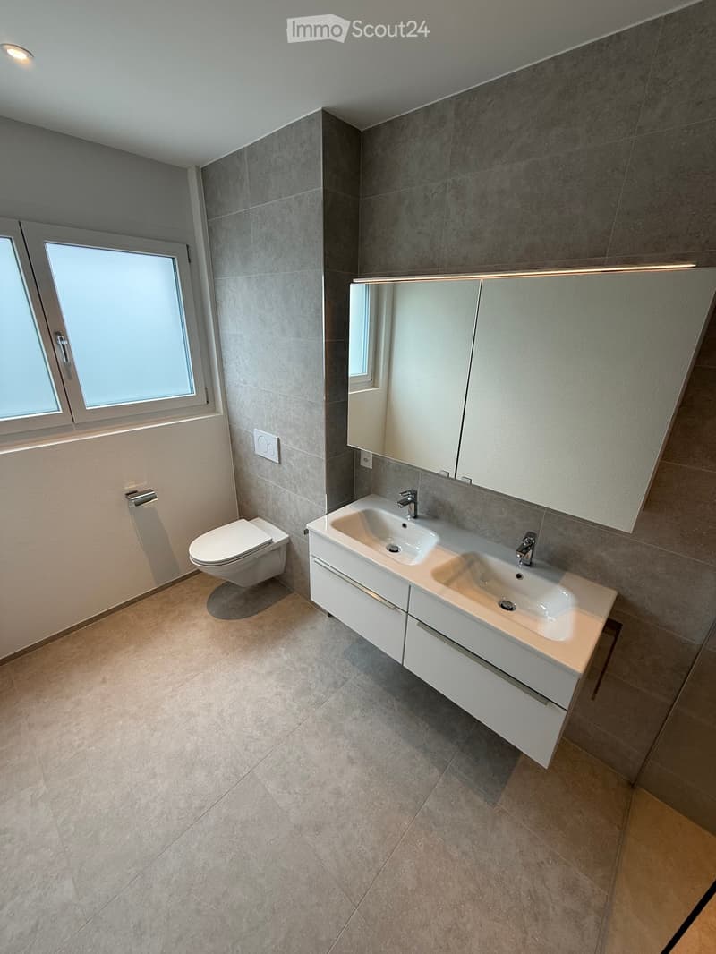 Modern bathroom with grey tiled walls, beige tiled floor, double sink vanity with white cabinets, toilet, and large mirror above the sink.