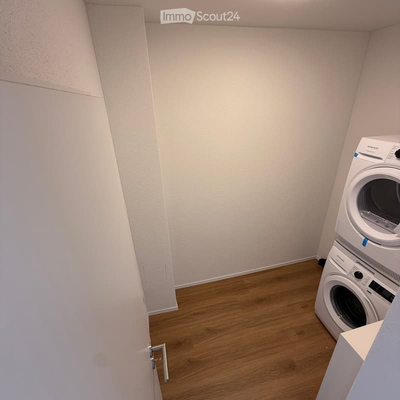 A small, unfurnished room with light-colored walls and wooden flooring, featuring a stacked washing machine and dryer unit.