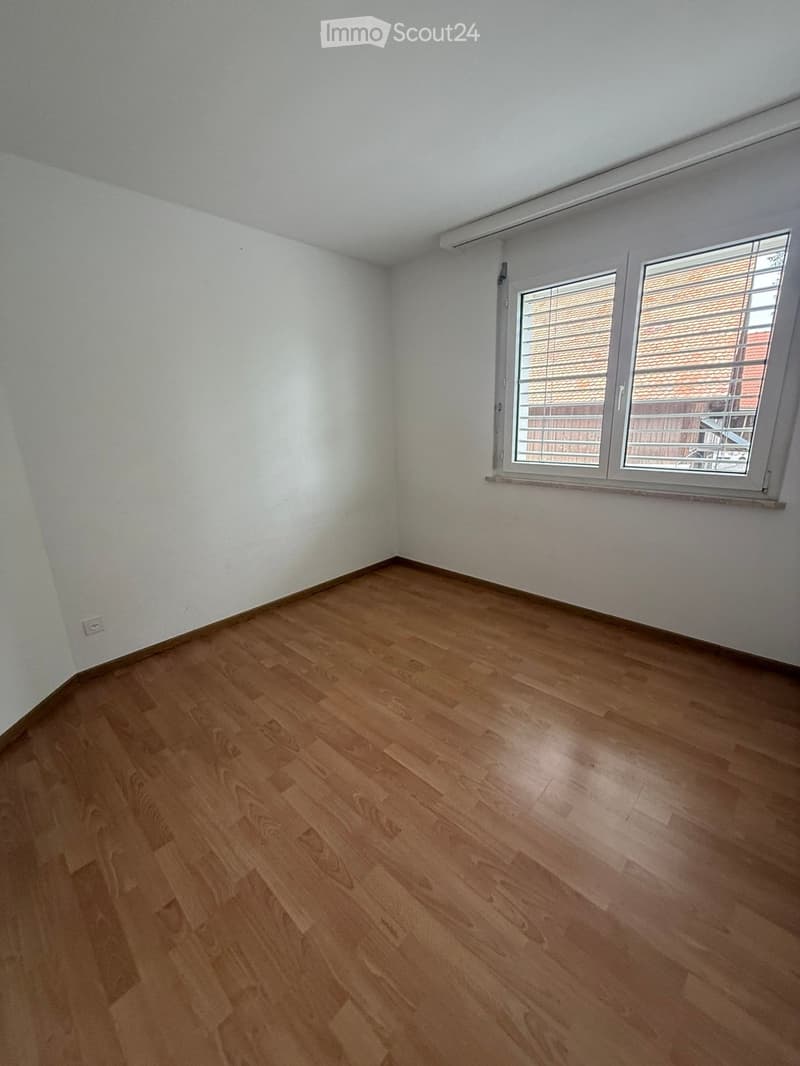 Empty room with wooden flooring and white walls, featuring a large window with blinds and a visible electrical outlet on the wall.