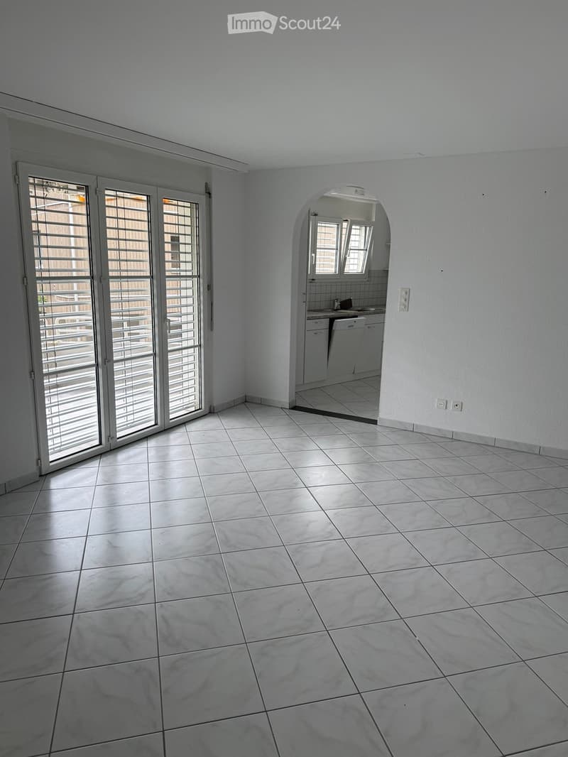 Empty room with tiled floor, white walls, large sliding glass doors leading to a balcony, and an arched doorway to a small kitchen area with white cabinets and a dishwasher.