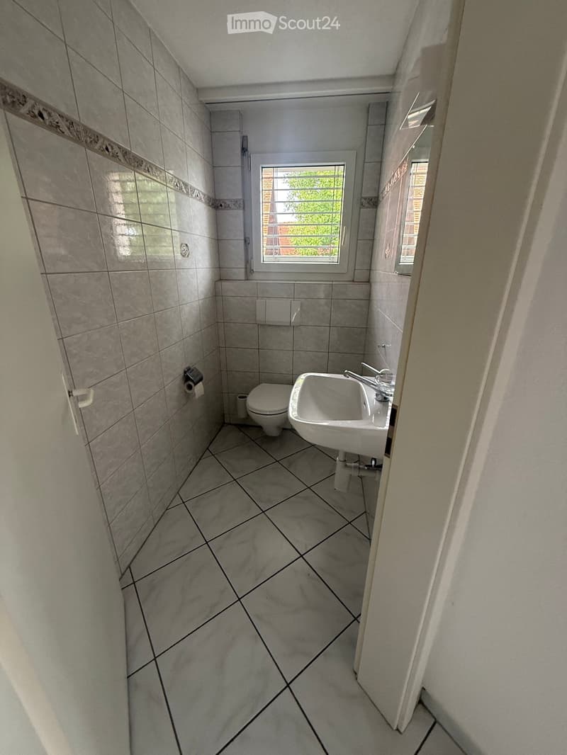 Small bathroom with grey tiled walls and floor, featuring a toilet, sink, and window with a view of greenery.