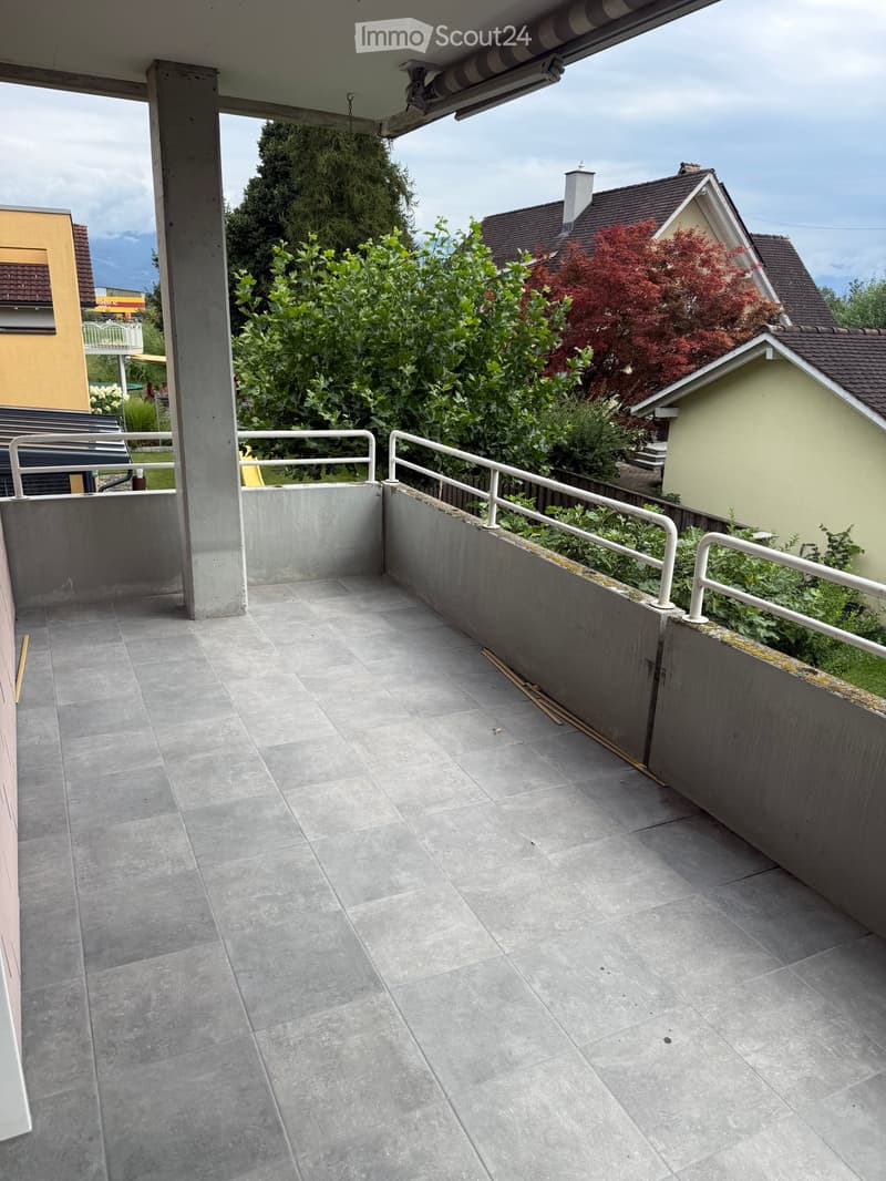 Balcony with grey tiled floor, white metal railing, and surrounding greenery.