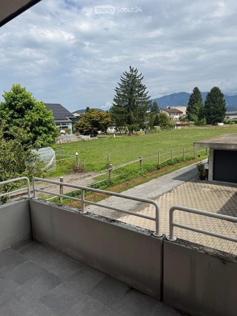Balcony with tiled floor, white metal railing, overlooking a fenced green field, trees, and distant mountains.