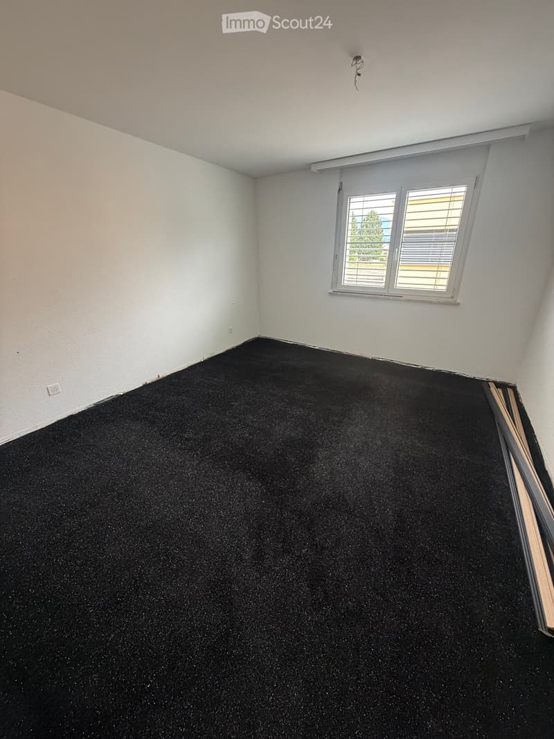 Empty room with black carpet flooring, white walls, and a window with blinds. No furniture or equipment present.