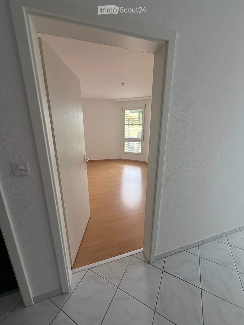 Empty room with wooden flooring and white walls, featuring a window with blinds and a door leading to another area.