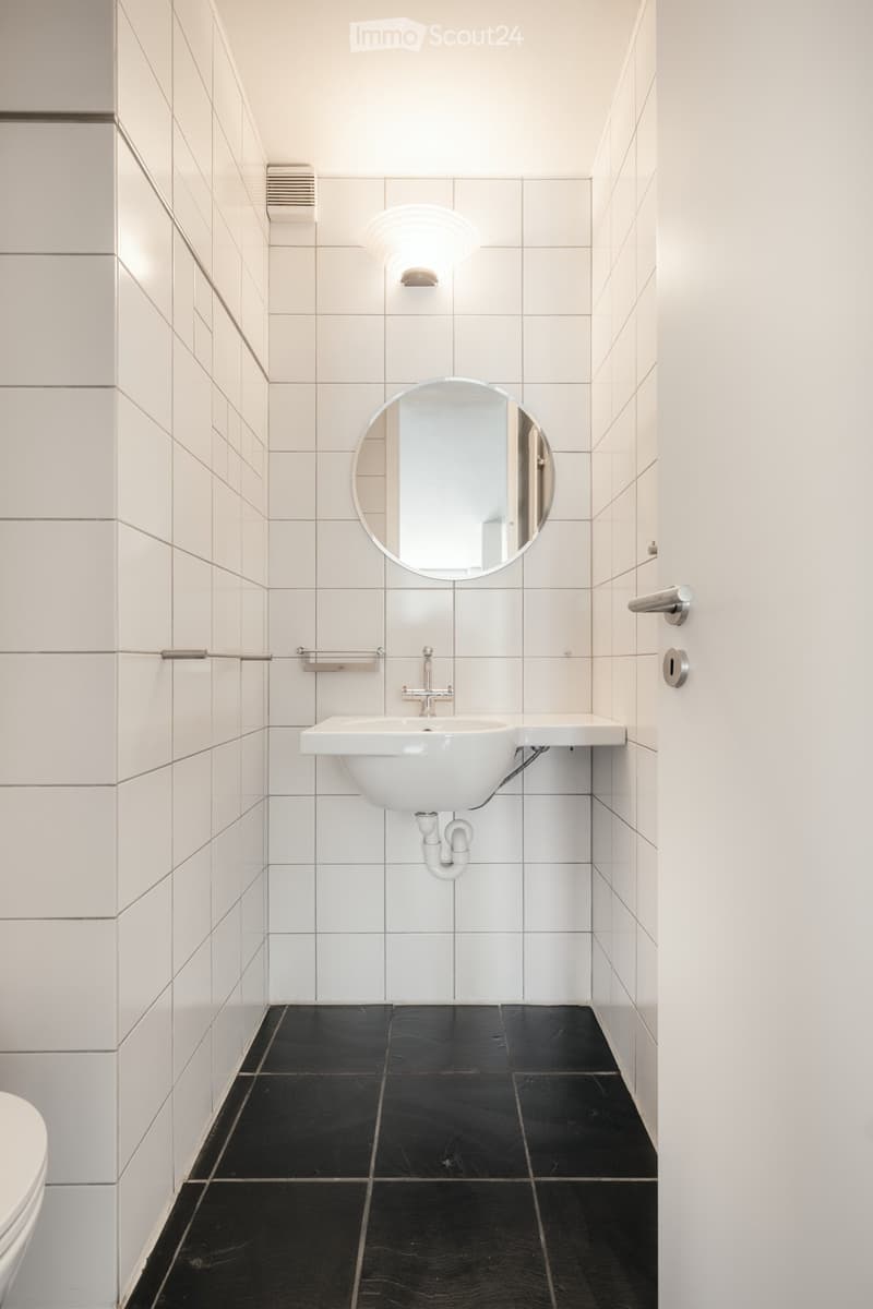 Modern bathroom with white tiled walls, black tiled floor, white sink, chrome faucet, round mirror, and toilet.
