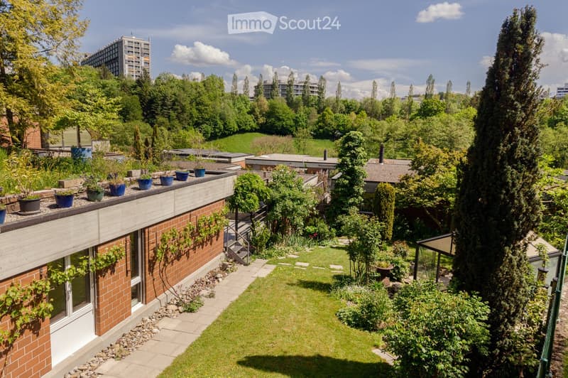 A well-maintained garden with a variety of plants, trees, and a small lawn. The garden features a brick wall, a staircase leading to a rooftop area, and several potted plants on the rooftop.