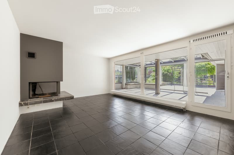 Modern living room with dark tiled flooring, large glass walls leading to an outdoor area, and a prominent fireplace with a dark surround.
