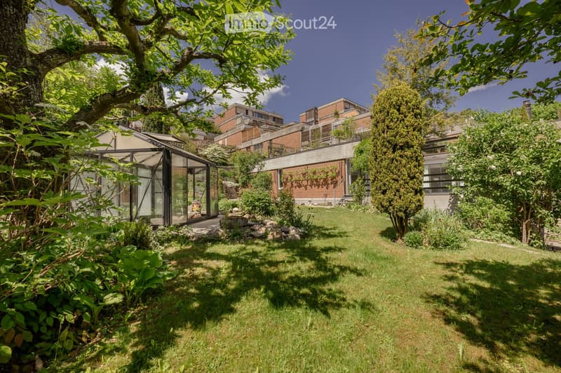 Well-maintained garden with a lawn, various trees, shrubs, and a greenhouse structure. The area is surrounded by residential buildings.