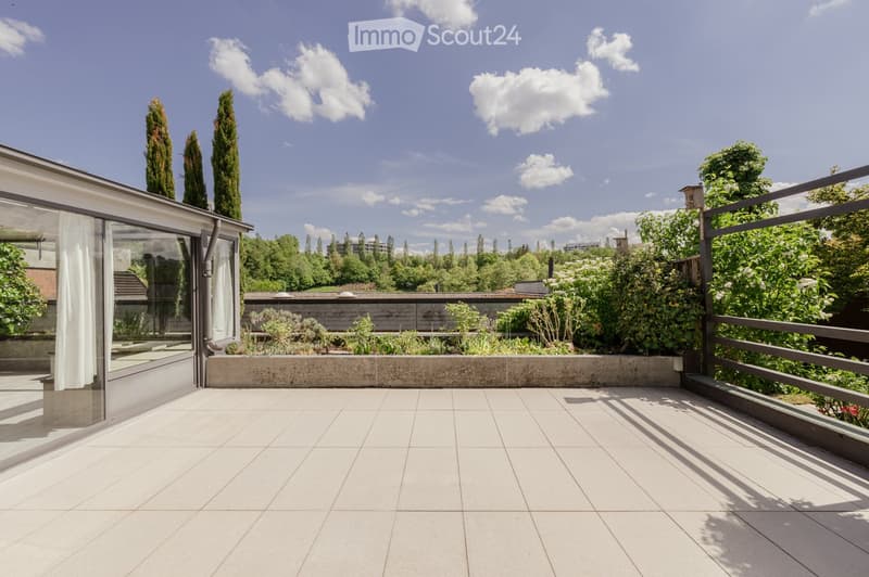 A modern rooftop terrace with white tiled flooring, glass walls, and a metal railing, surrounded by greenery and trees.