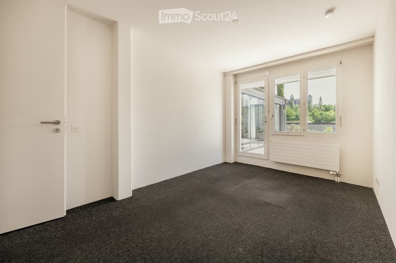 Empty room with white walls, grey carpet, large windows, and a sliding glass door leading to an outdoor area.