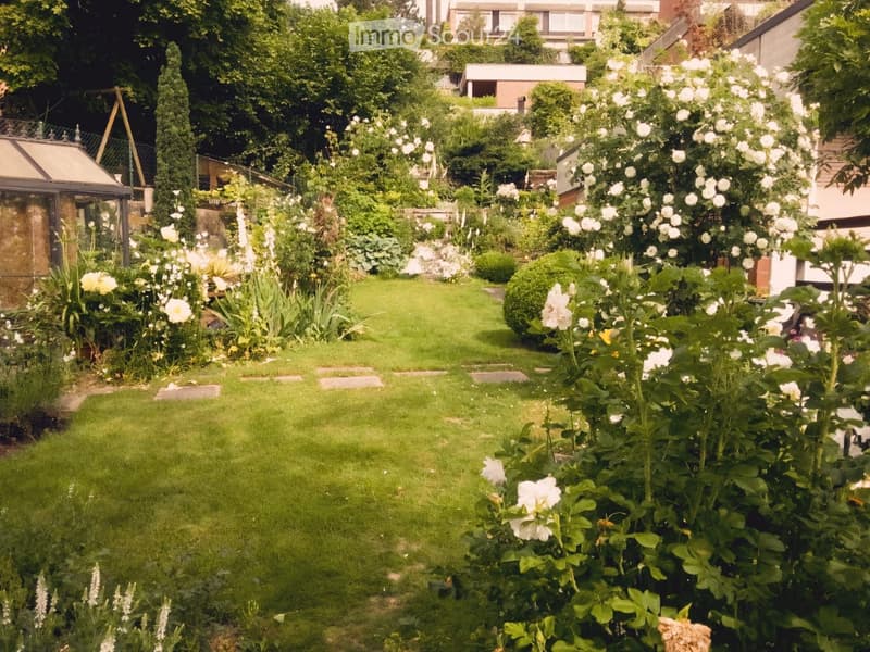 Well-maintained garden with a central lawn, surrounded by various flowering plants, shrubs, and trees. Features stepping stones leading through the garden and a small greenhouse on the left side.