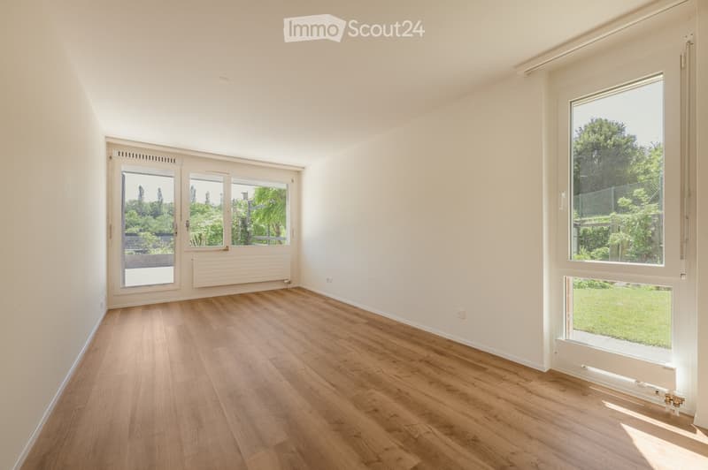 Empty room with light-colored walls, wooden flooring, and large windows offering views of greenery outside. No furniture or equipment present.