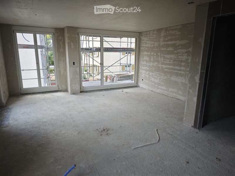 Empty room with unfinished concrete floor and walls, large glass doors leading to a balcony area with construction materials visible outside.