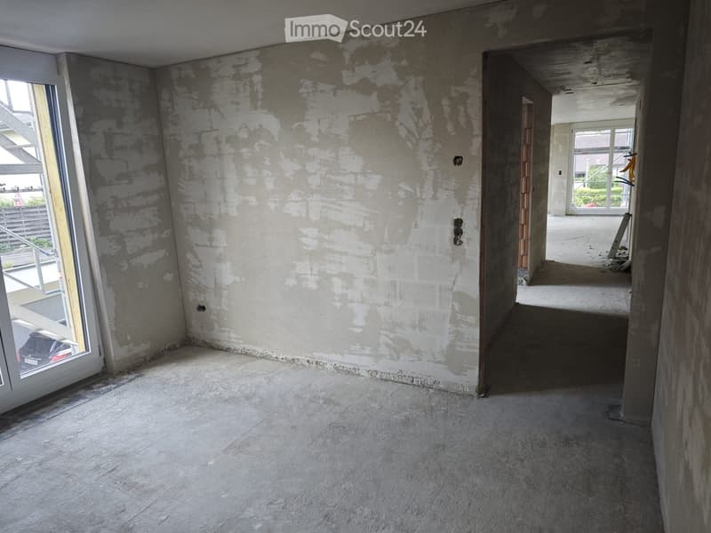 Empty room with unfinished concrete walls and floor, large window on one side, and an open doorway leading to another room.