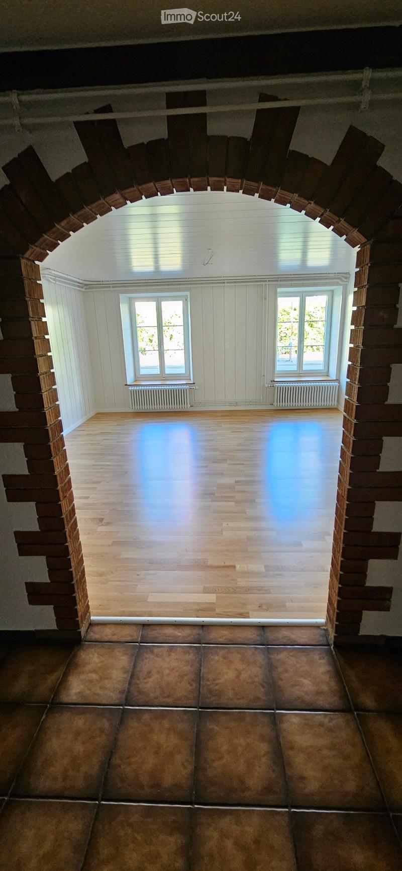 An empty room with wooden flooring and white walls, featuring two windows and radiators, with a raised wooden platform in the center.