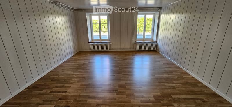 Empty room with wooden flooring, white paneled walls, and two windows with radiators beneath them.