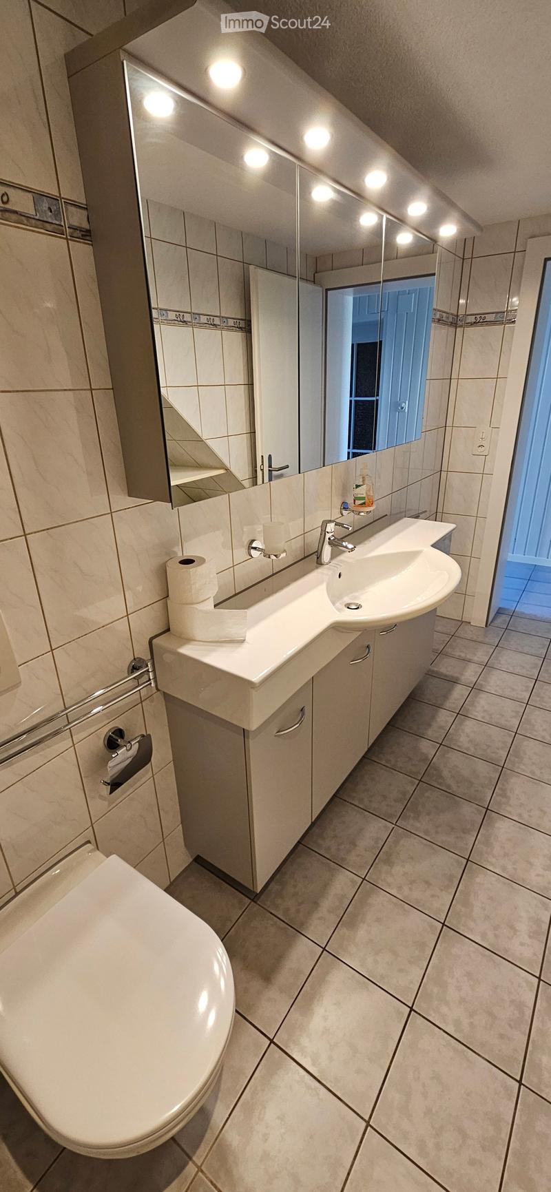 Modern bathroom with a white sink, toilet, large mirror, and built-in cabinet. The walls are tiled in beige, and the floor is tiled in light gray.