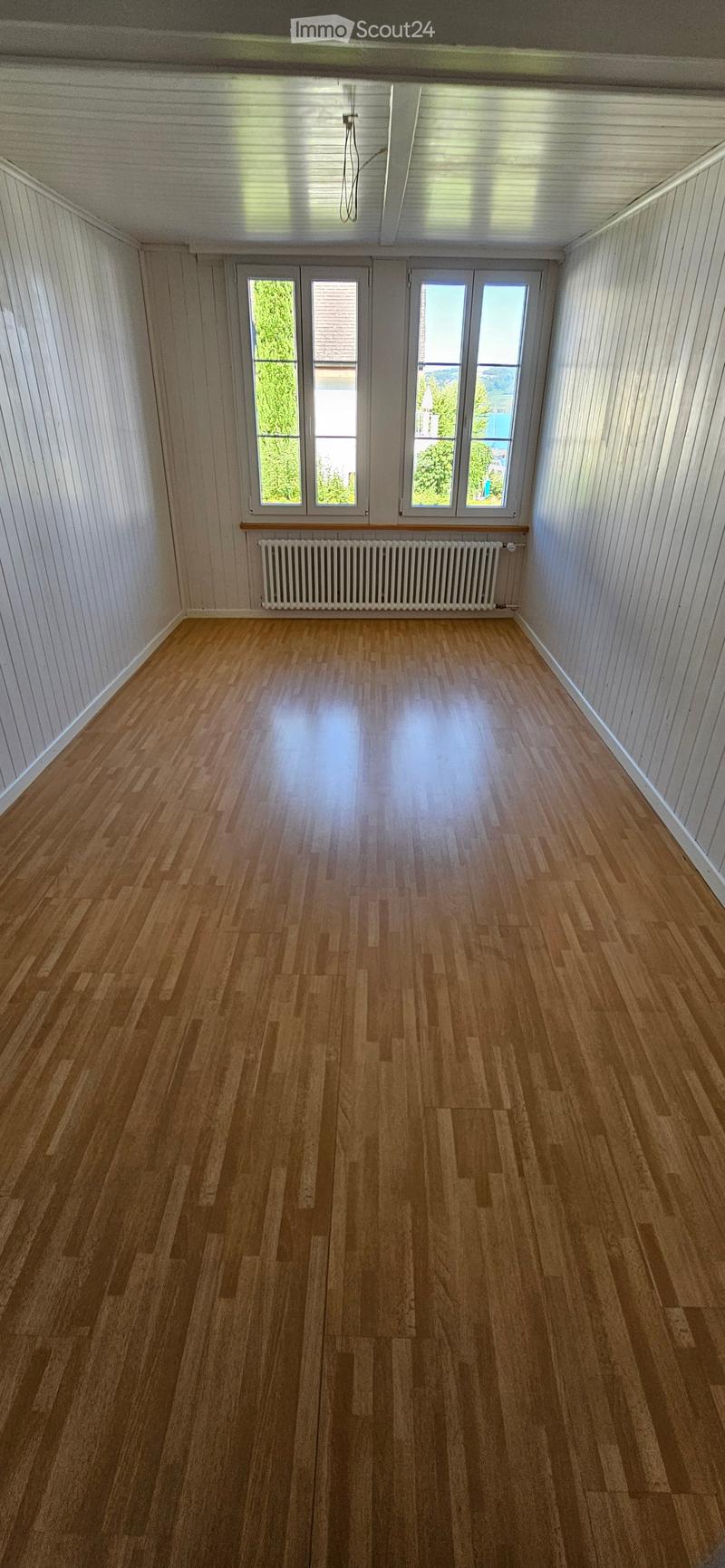 Empty room with wooden flooring, white paneled walls, and large windows with a radiator below them.