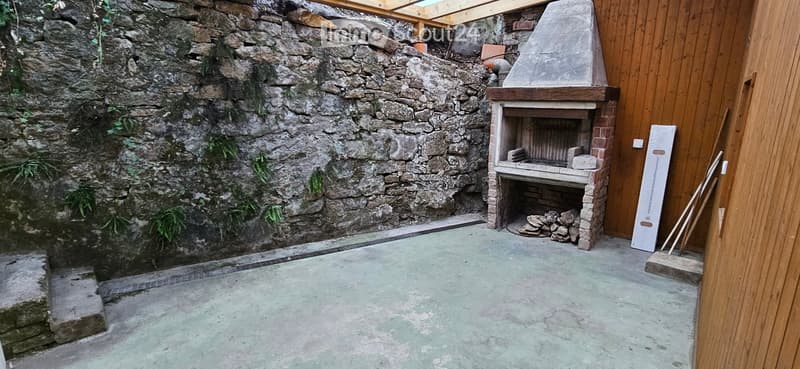Outdoor space with a stone wall, wooden fence, and a brick barbecue with a chimney. Concrete floor and some greenery on the wall.