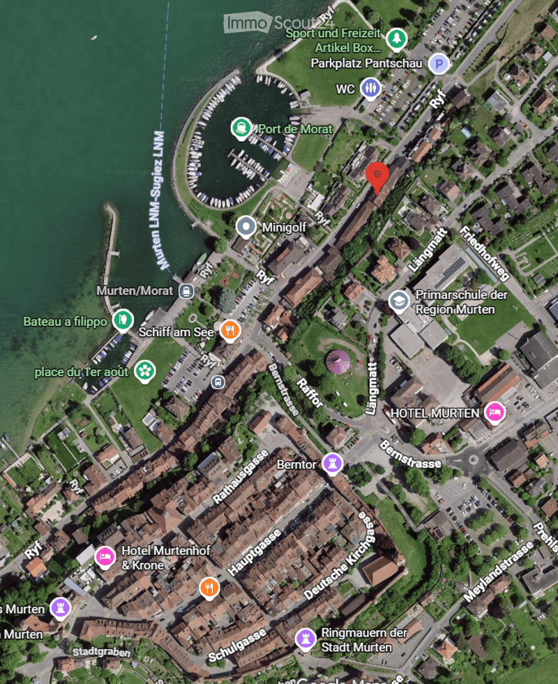 Aerial view of a town with buildings, streets, and a waterfront area with a marina. Visible landmarks include a hotel, school, and recreational areas.