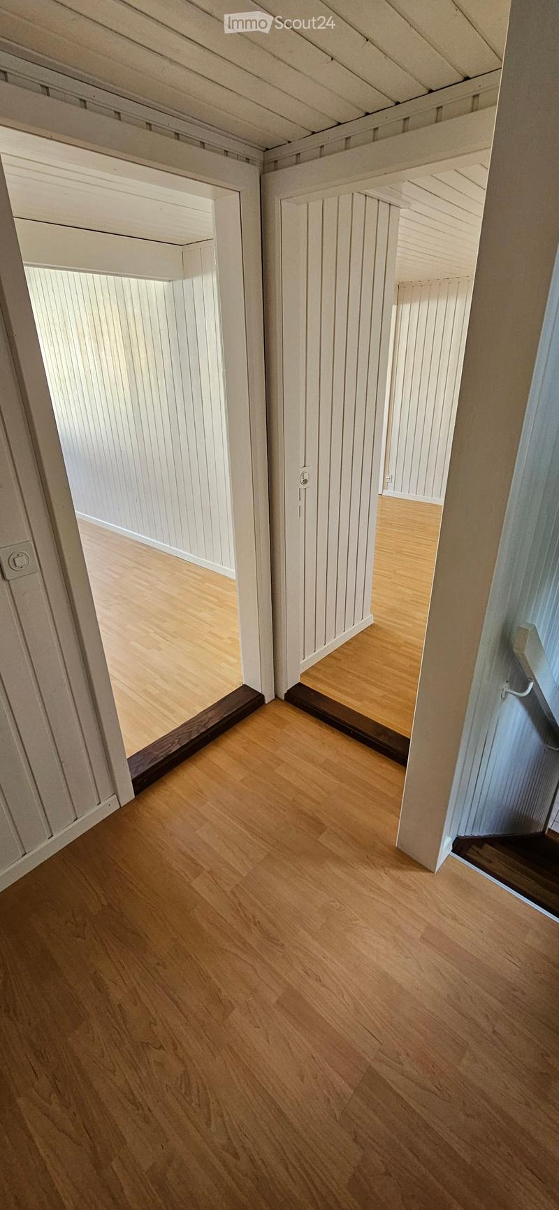 Empty hallway with light wooden flooring and white paneled walls, featuring open doorways leading to adjacent rooms.