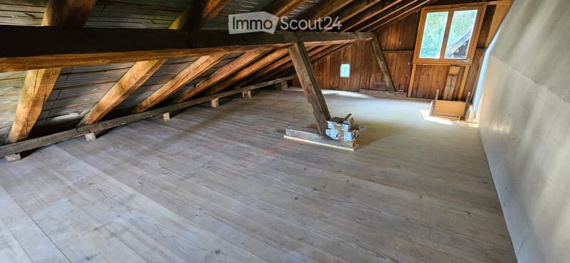 An attic space with wooden beams, exposed wooden floor, and a small window letting in natural light. The room is empty, with no furniture or equipment.
