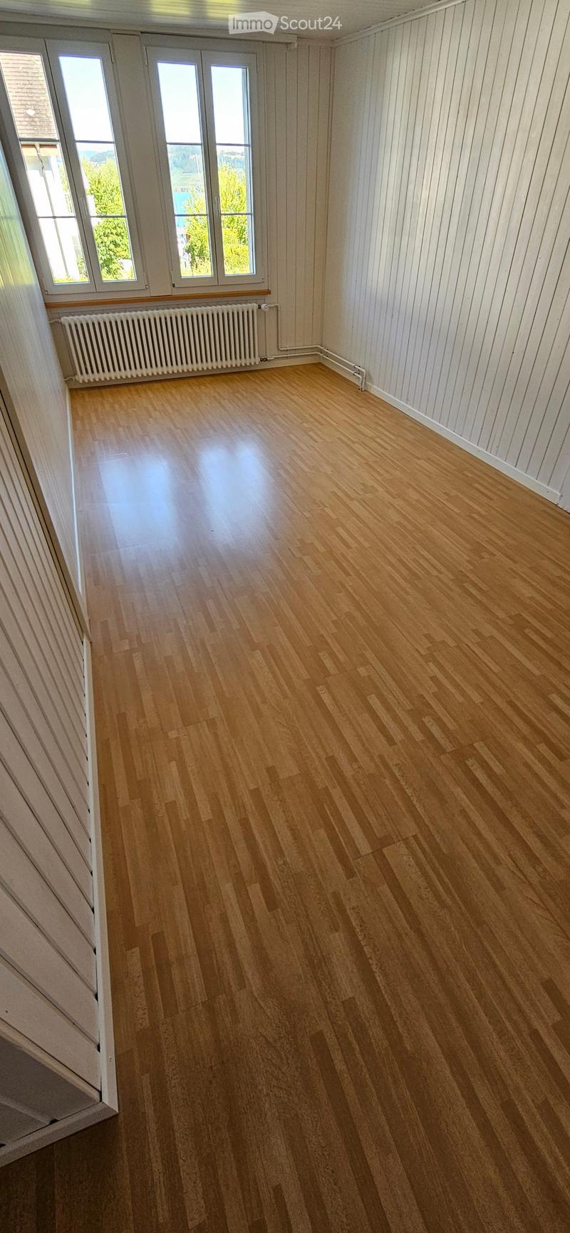 Empty room with wooden flooring, white walls, and large windows with white frames. A radiator is visible along one wall.