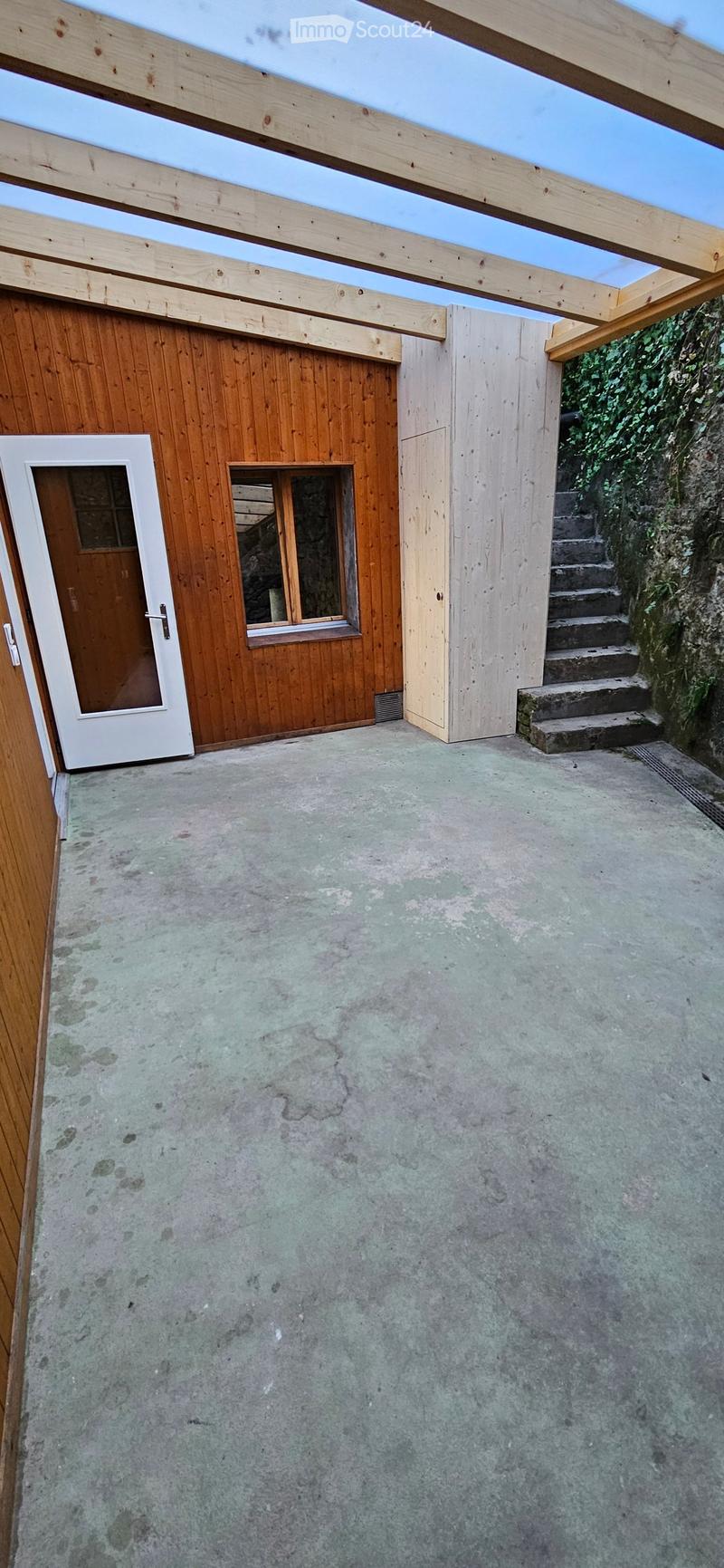 An outdoor patio area with a concrete floor, wooden walls, a small window, a white door, and a set of concrete steps leading up to a wooden structure with a slanted roof.