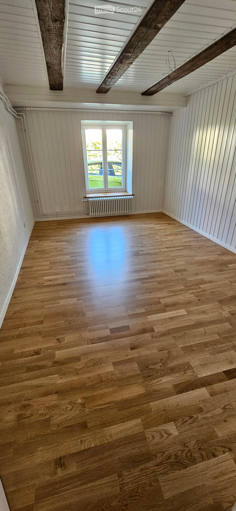 Empty room with wooden flooring, white paneled walls, and a single window with a radiator below it. The ceiling features exposed wooden beams and a skylight.