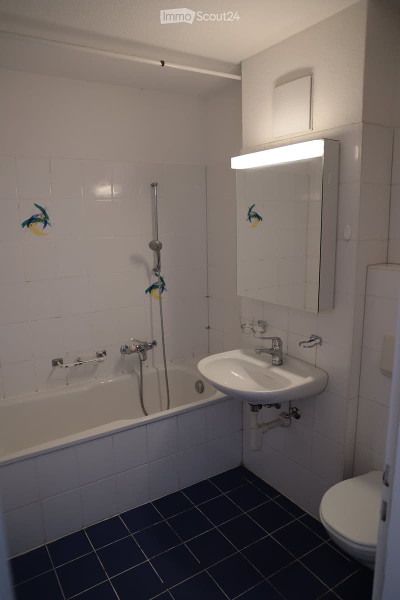 Bathroom with a bathtub, sink, mirror, and toilet. The walls are tiled with light-colored tiles, and the floor is dark blue tiles.