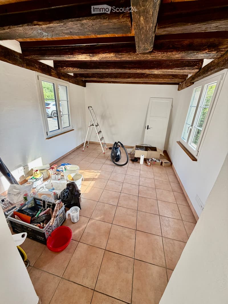 Empty room with tiled floor, white walls, wooden beams on the ceiling, windows, and a door. Various tools and materials are scattered around, indicating ongoing renovation or cleaning.