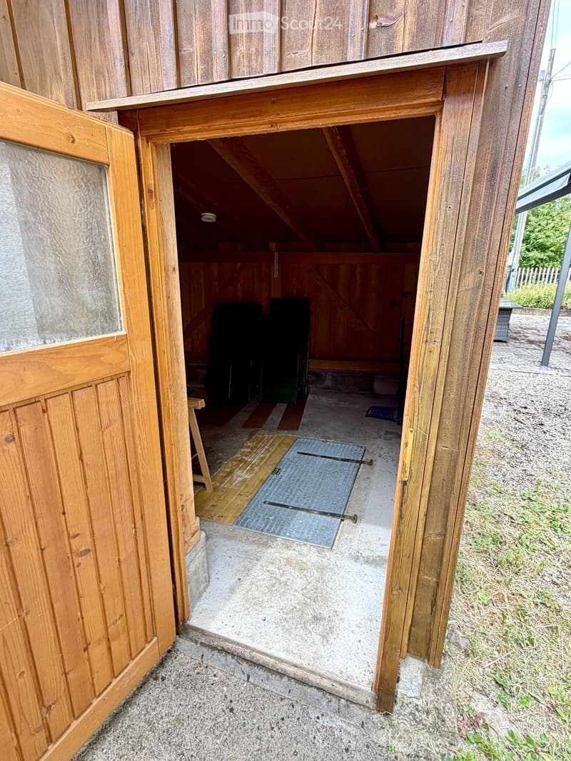 An open wooden shed with a concrete floor, featuring a small ramp leading up to a raised wooden platform. The shed contains a green container and a blue mat on the floor.