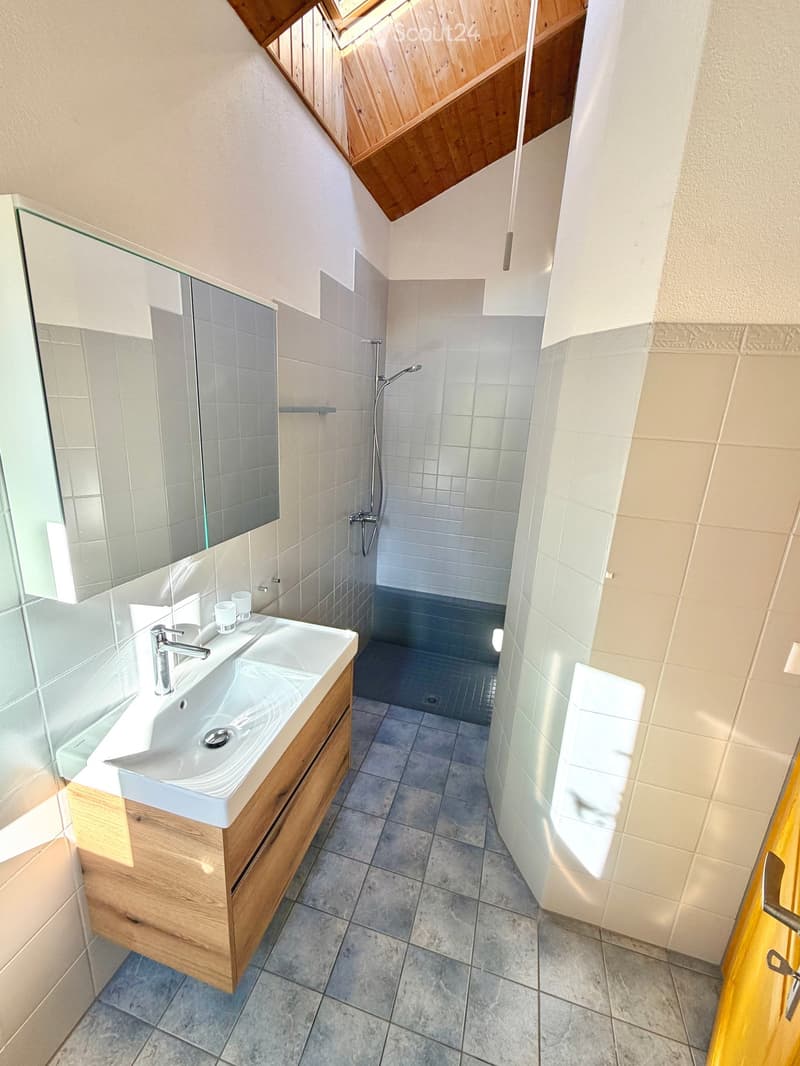 Modern bathroom with a wooden vanity, sink, mirror, and a separate shower area with a glass enclosure. The space features light-colored tiles and a skylight providing natural light.