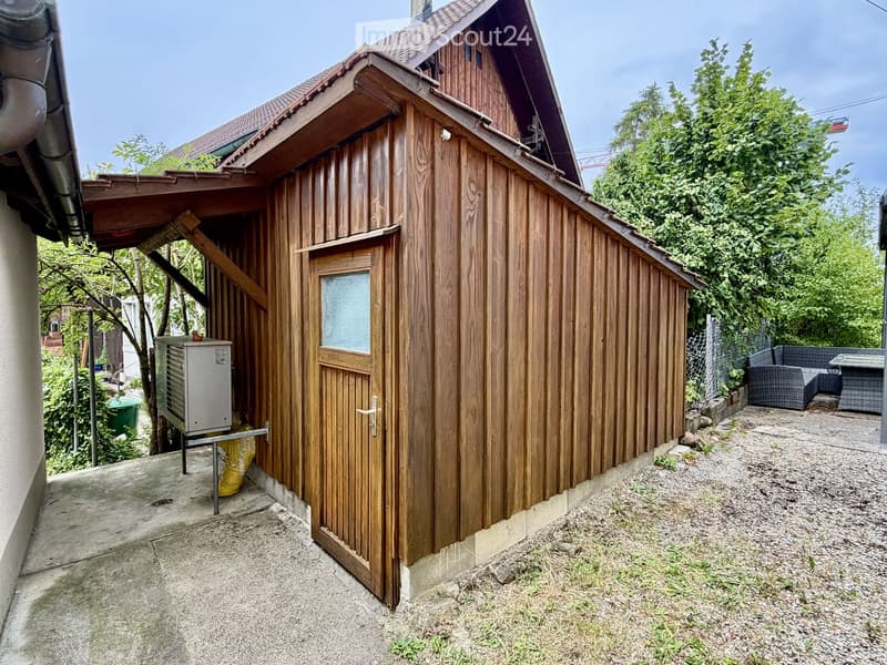 A small wooden shed with a slanted roof, a door with a frosted glass panel, and an air conditioning unit placed next to it. The shed is situated on a concrete patio with gravel ground and surrounded by greenery.