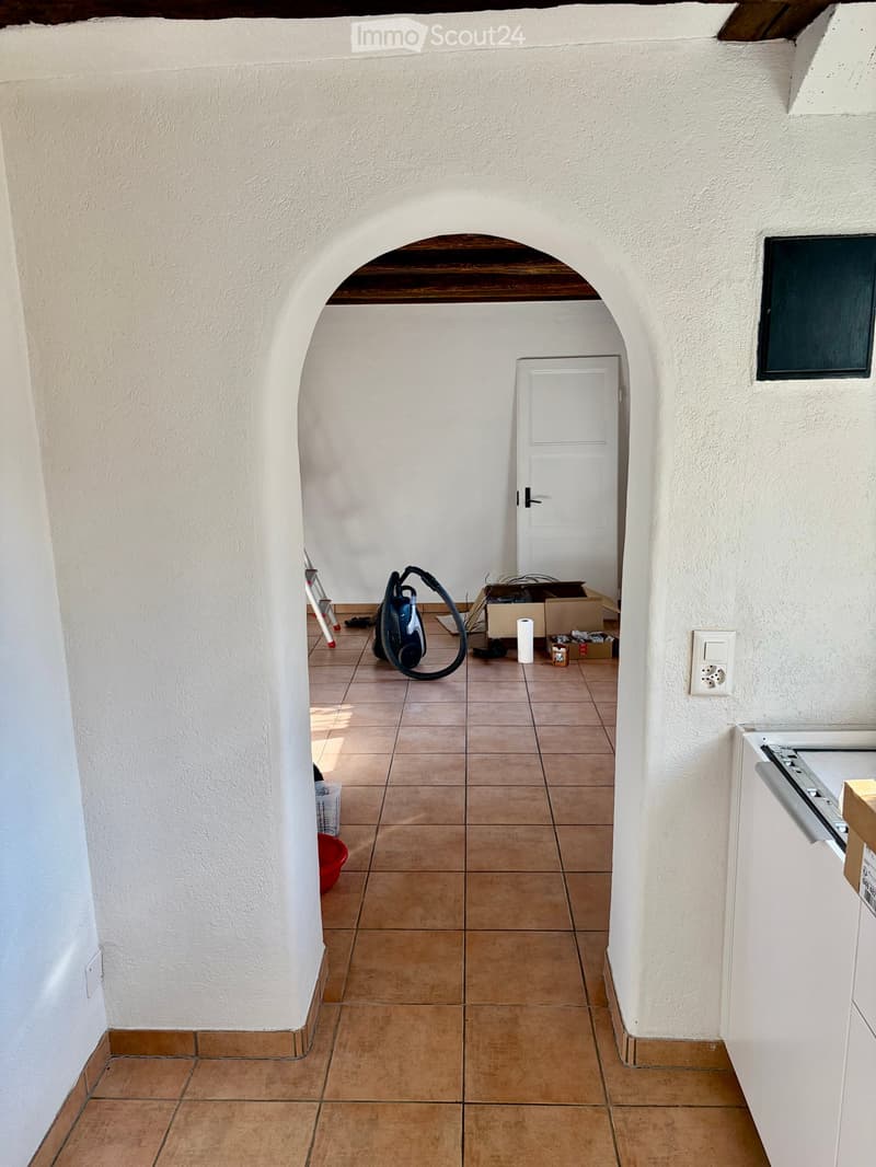 Empty hallway with tiled floor, white walls, and an arched doorway leading to another room. There are some cleaning supplies and boxes in the adjacent room.
